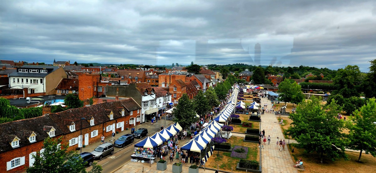A high-angle, wide view of Stratford-upon-Avon on a cloudy day, showing a row of historic brick buildings with dark tiled roofs on the left and a long line of blue-and-white striped market stalls stretching down a pedestrian walkway. Lush green trees and the town's skyline are visible under a dramatic grey sky."