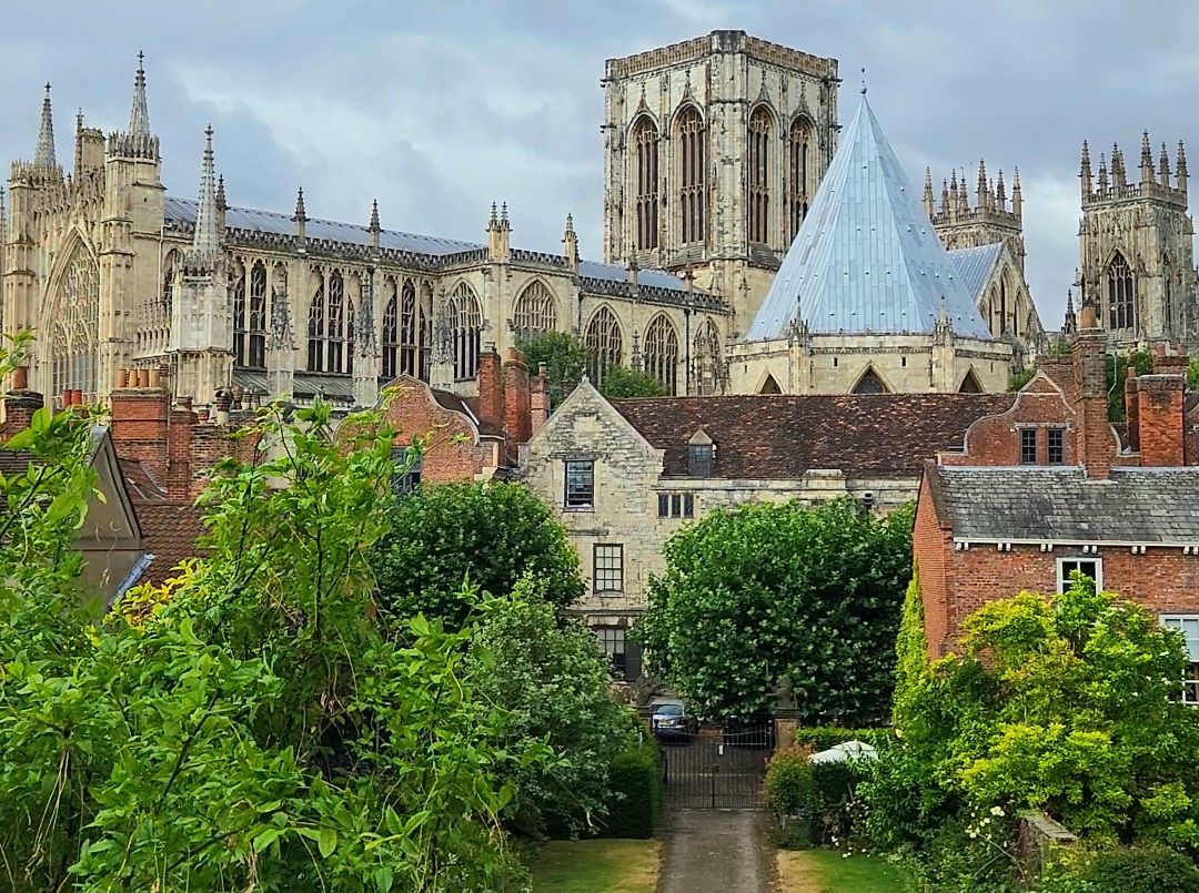 A stunning view of York Minster’s grand Gothic architecture and towers rising behind historic red-brick buildings. Lush green trees and a small stone path in the foreground frame the cathedral under a dramatic, overcast sky.